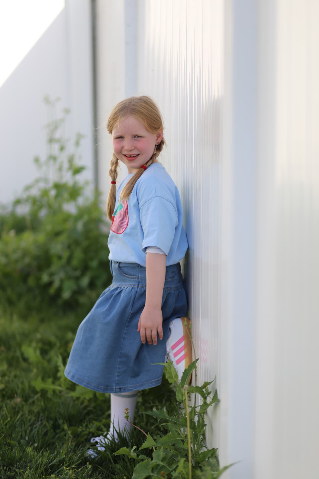 A girl in her strawberry t-shirt and denim skirt, resting against the wall with one foot up.