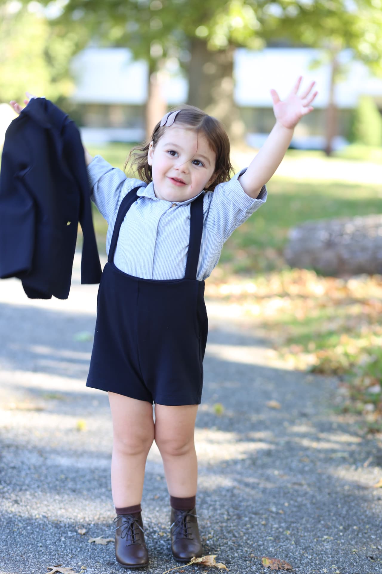 A little girl in suspenders throwing both arms up, jacket in one hand.