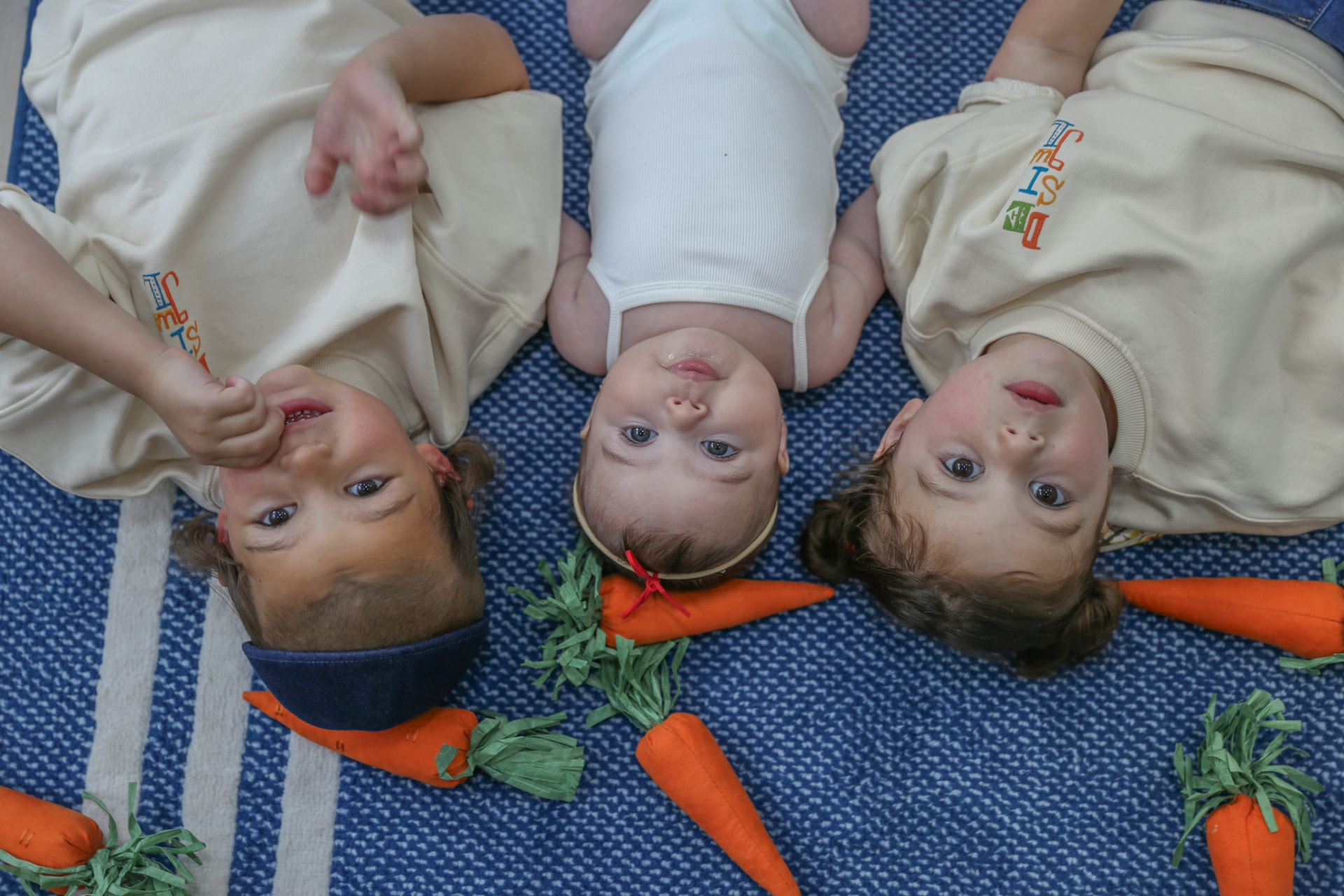 Three siblings lying upside down on the blue rug among felt carrots.