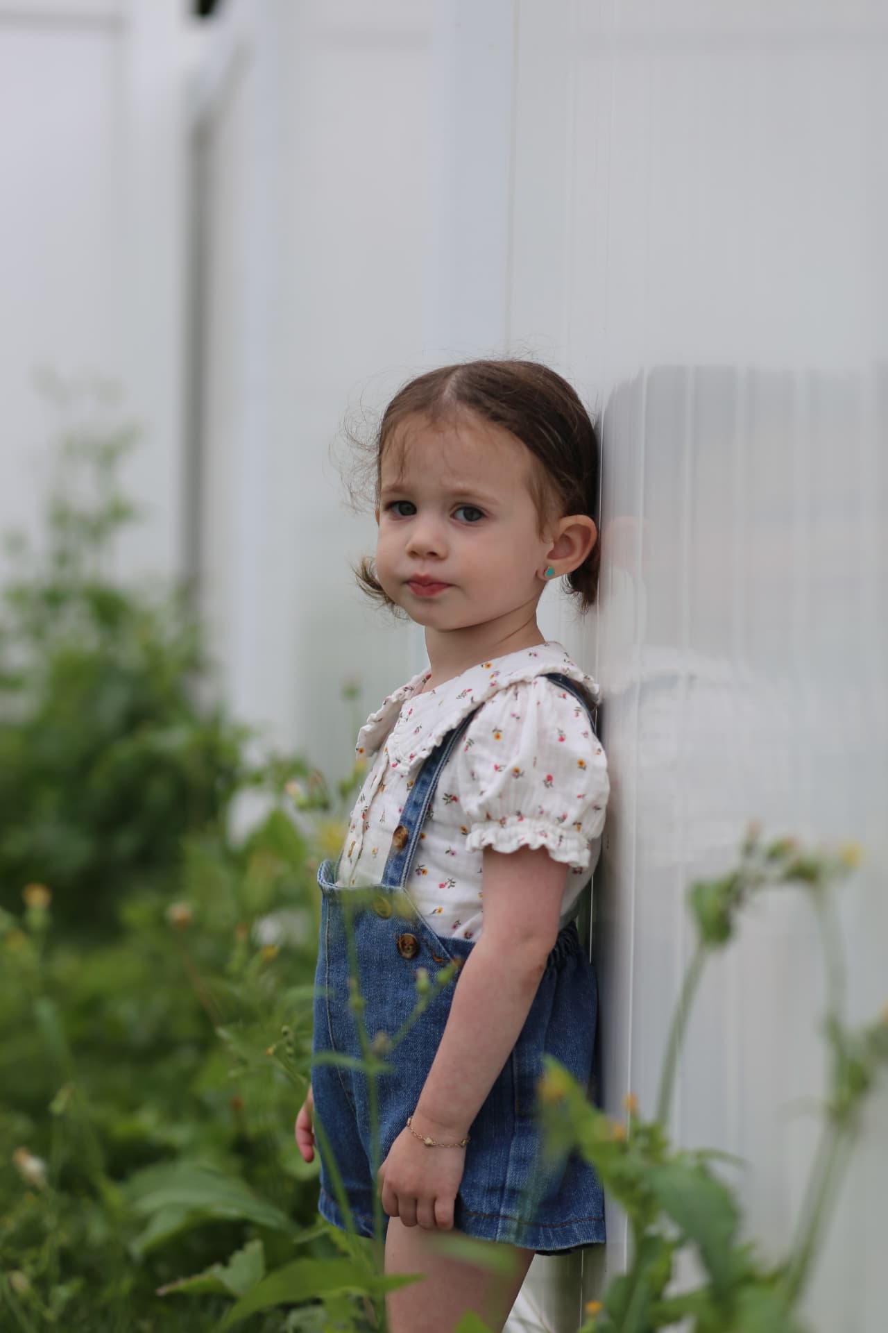A toddler girl in denim overalls and a flowered top, leaning against the white wall.