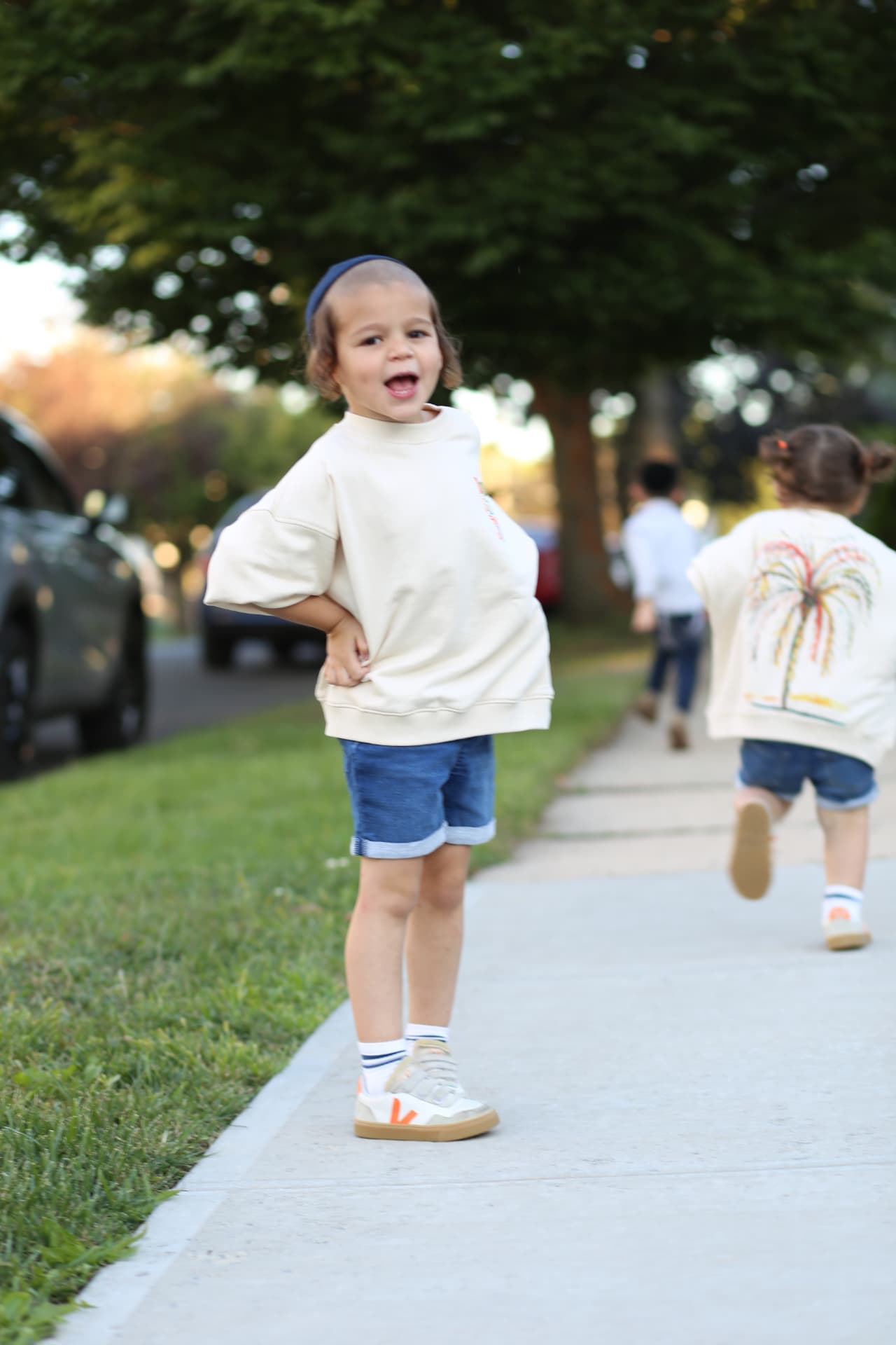 A boy striking a pose on the sidewalk while his sister runs ahead.