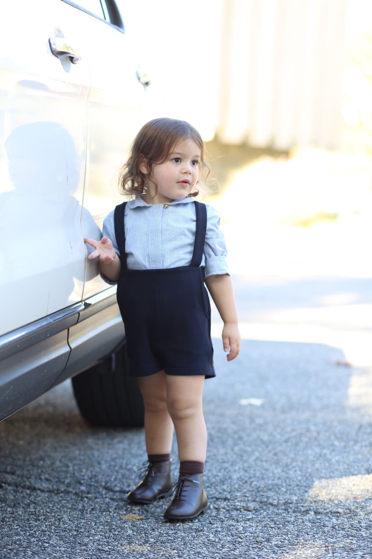 A toddler in suspenders and lace-up boots leaning on the car in the driveway.