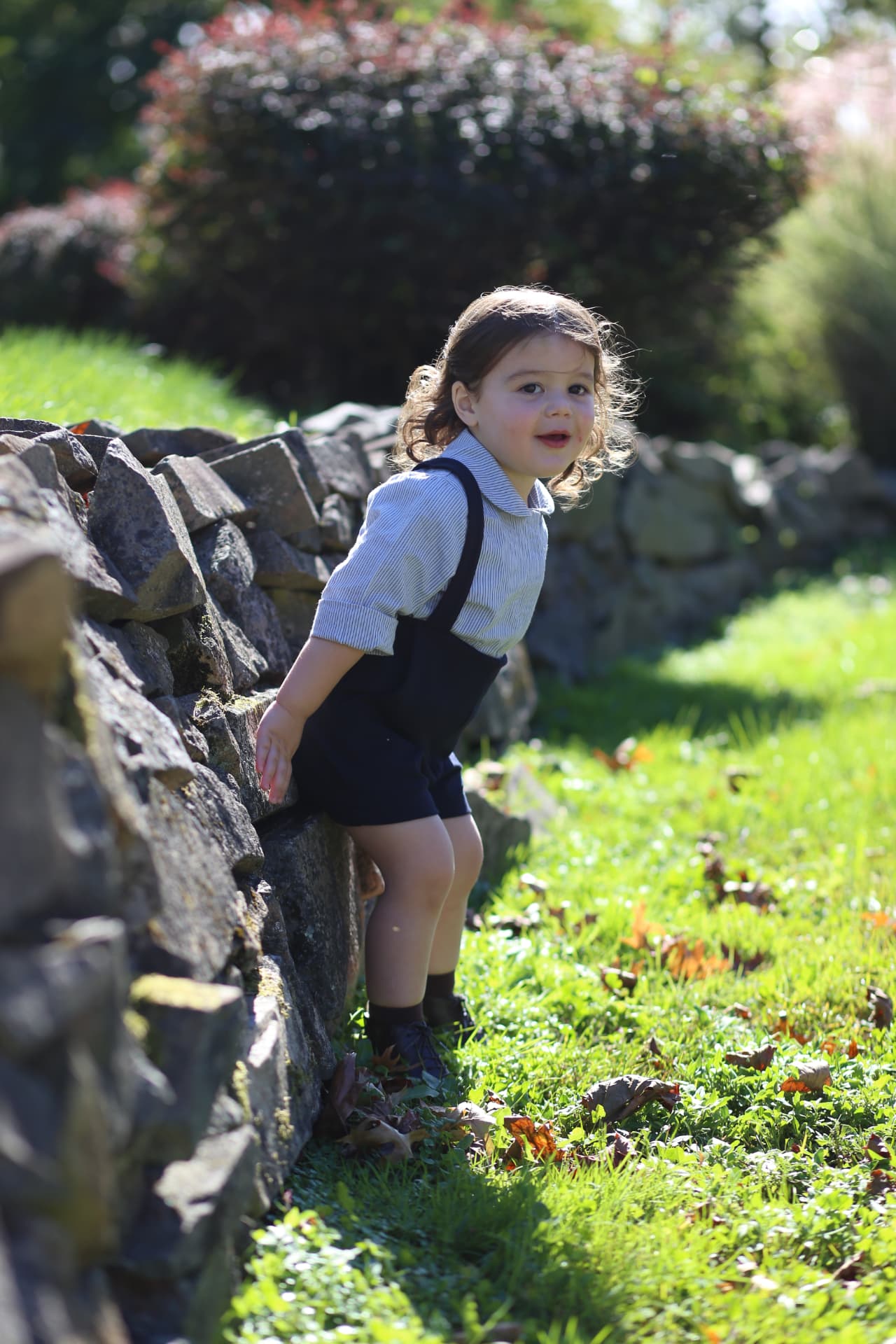 A curly-haired toddler climbing along a sunlit stone wall.