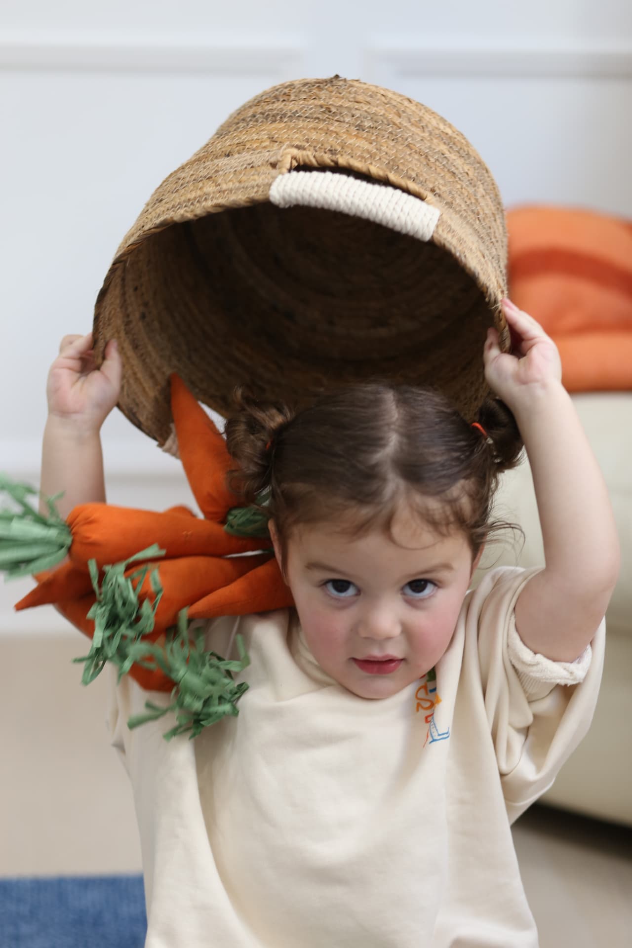 A toddler lifting a woven basket over her head, carrots tumbling out.