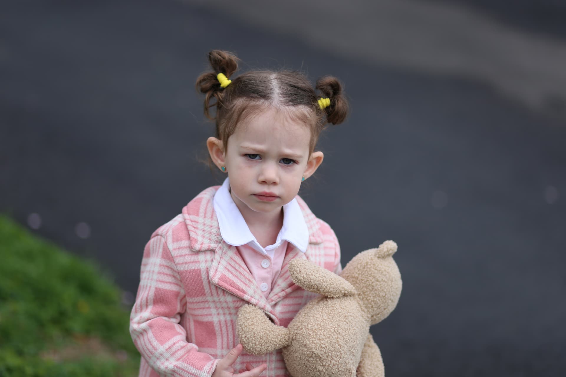 A girl in pink plaid, holding her bear close on the street.