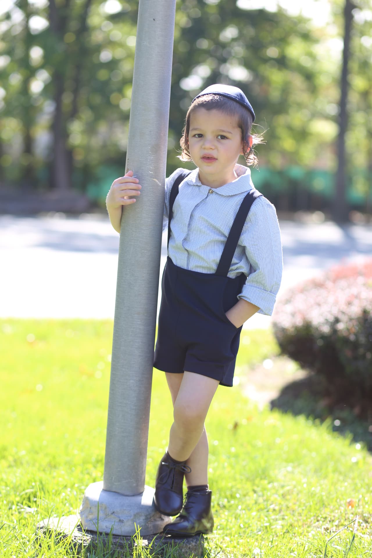 A boy in suspenders leaning against the pole, hand in his pocket.