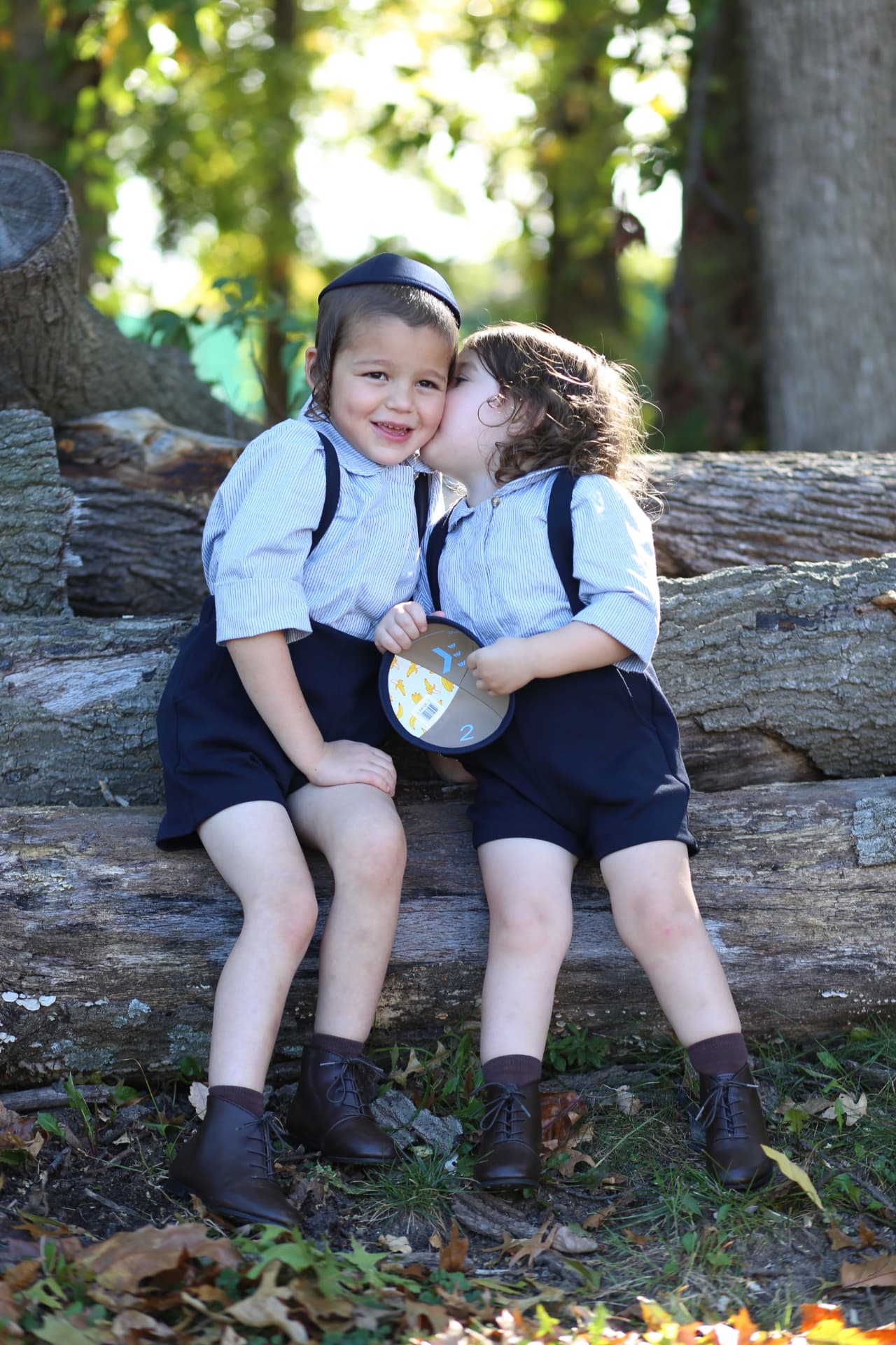 Little sister kissing her brother on the log pile, matching suspenders.