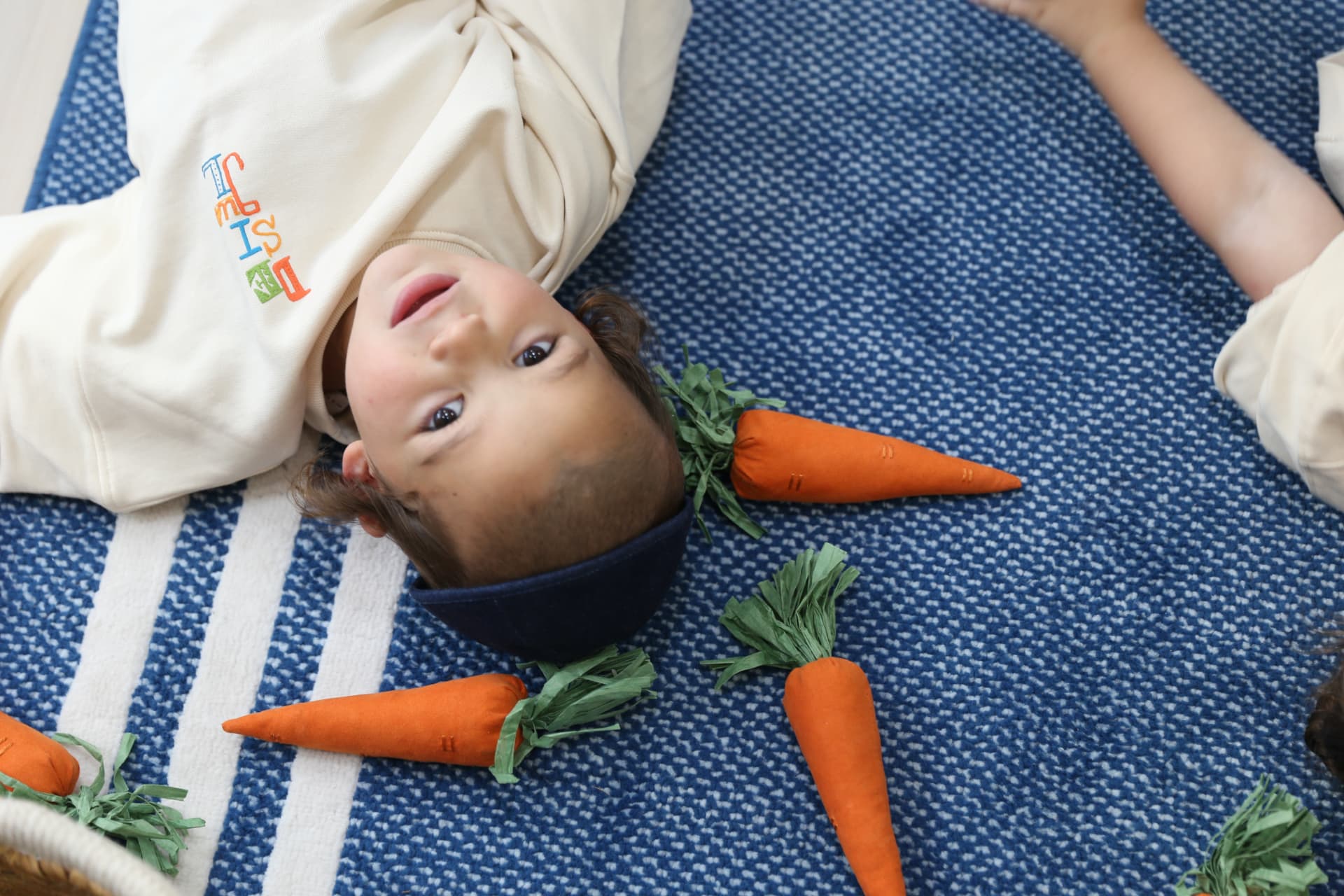A boy lying among plush carrots on the blue rug, looking up.