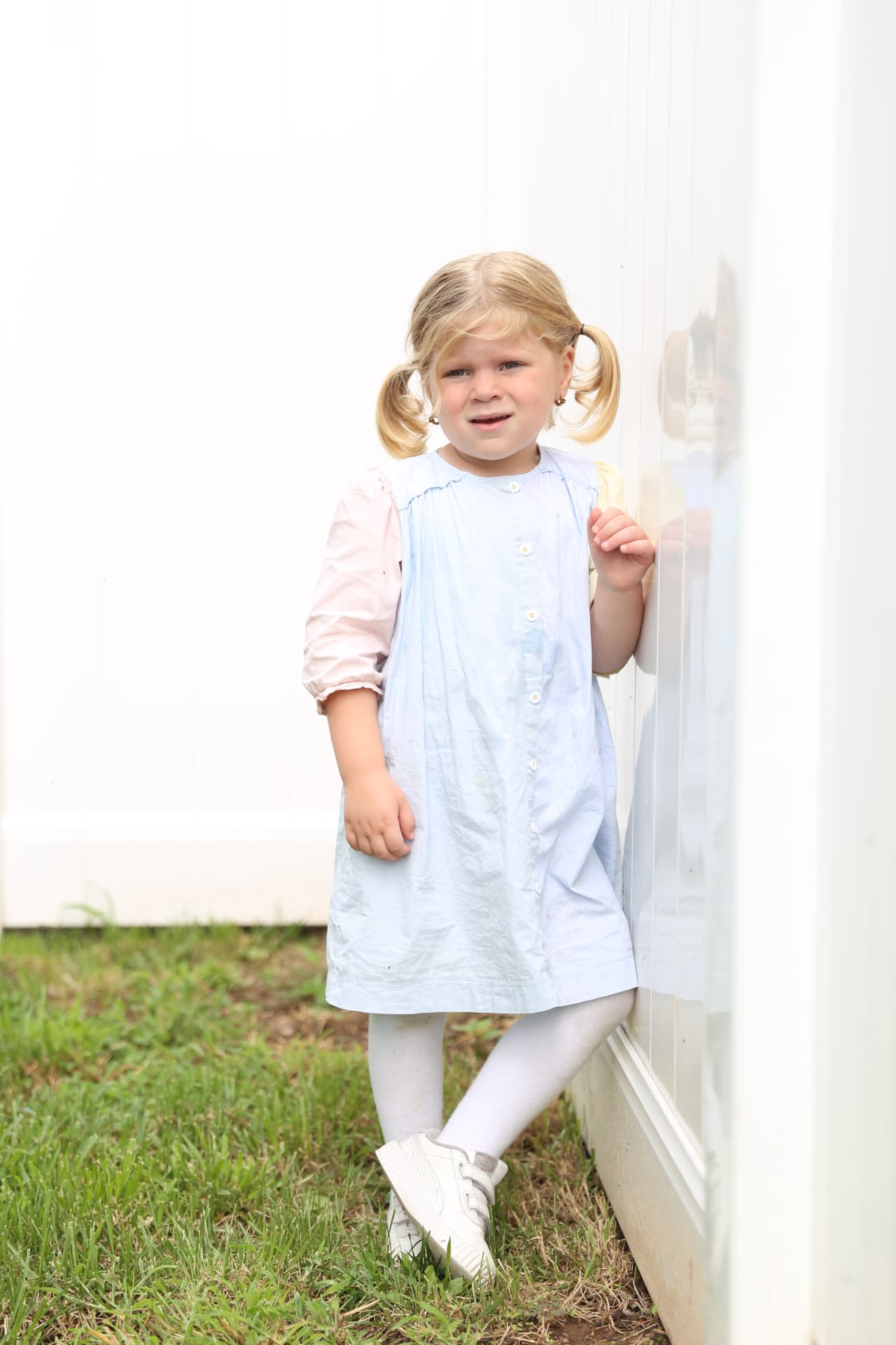 A little girl in pigtails standing in the grass by the white wall.