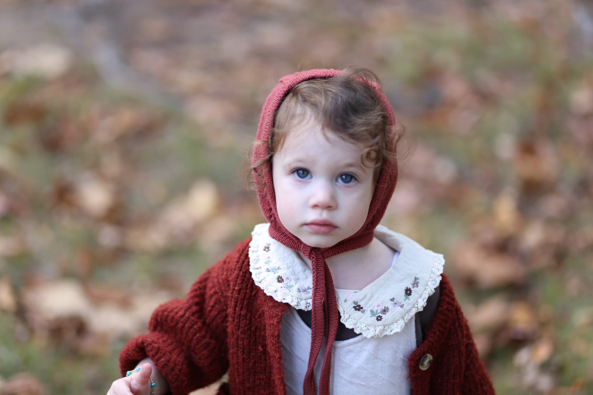 A little girl in a knit bonnet among fall leaves.