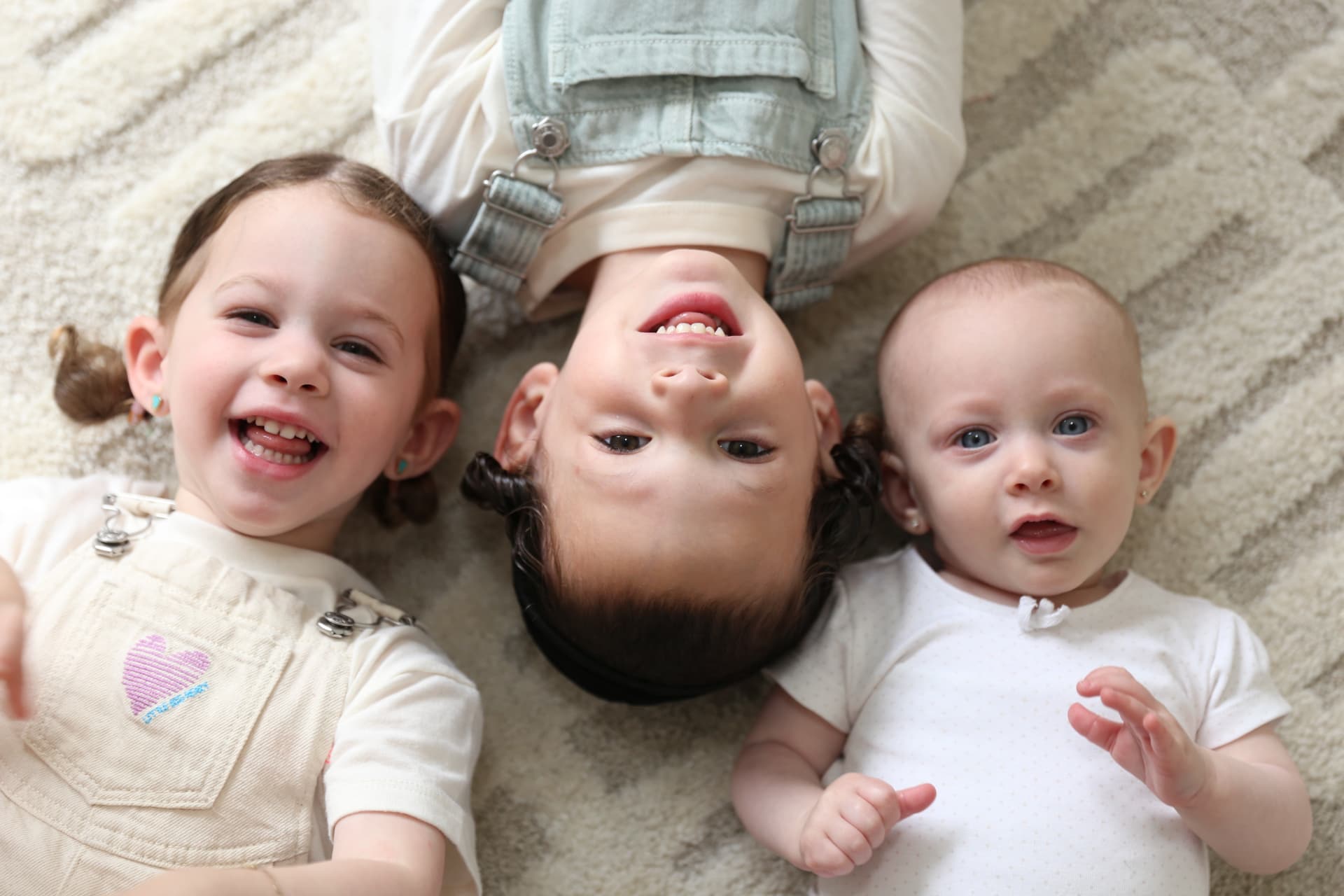 Three siblings lying head to head on the rug, two of them laughing.