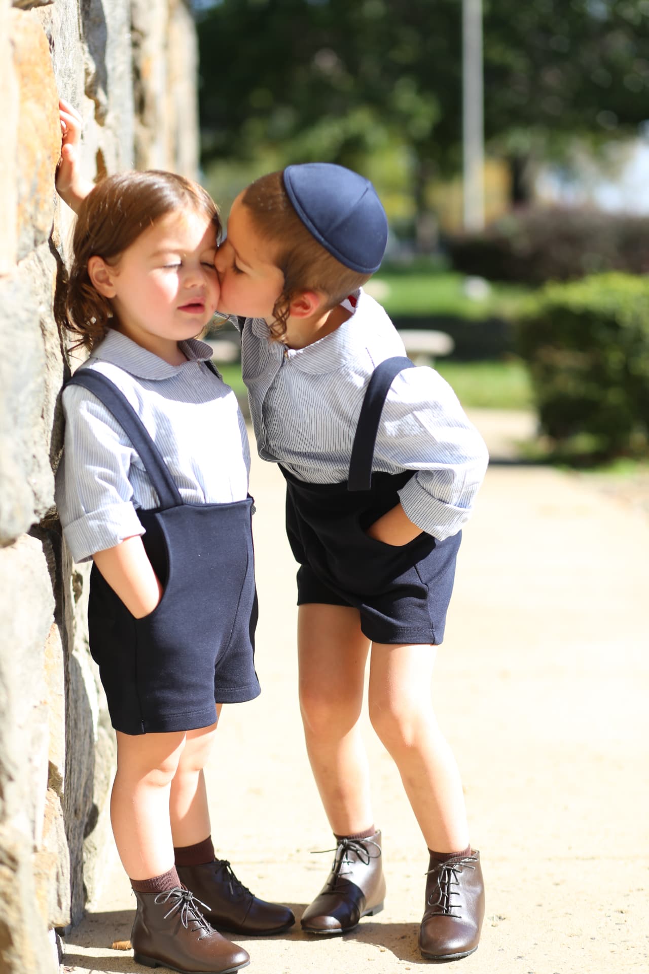 A big brother leaning in to kiss his little one by the stone wall.