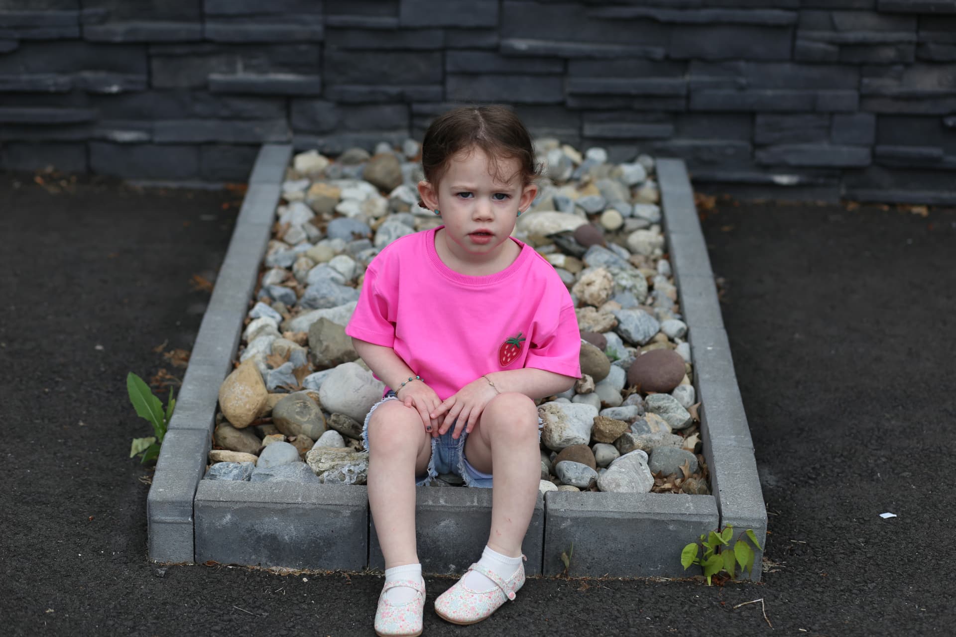 A little girl in pink sitting on the stone edge of the pebble bed.