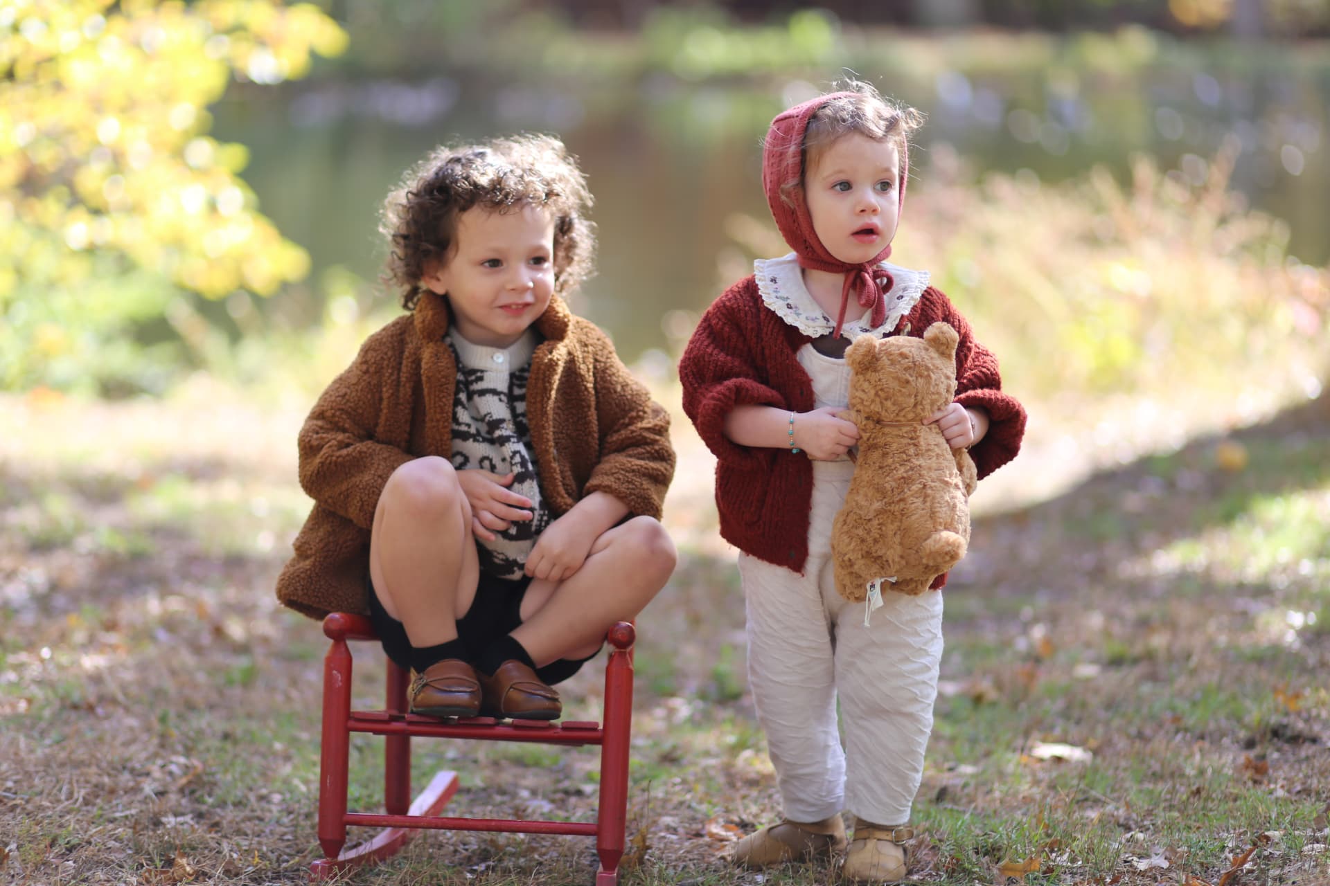 A curly-haired boy on a red rocking chair beside his sister holding her teddy bear.