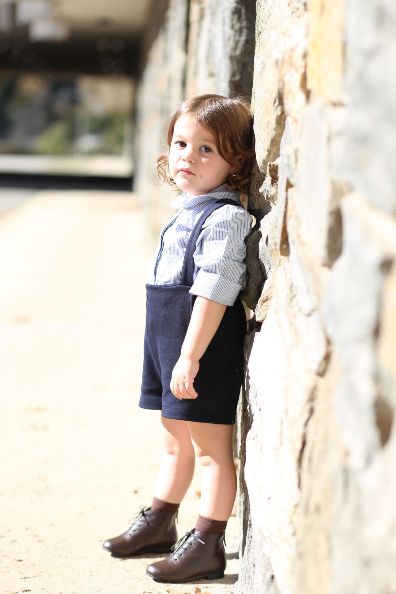 A girl in navy overalls resting her curls against the old stones, eyes on the camera.