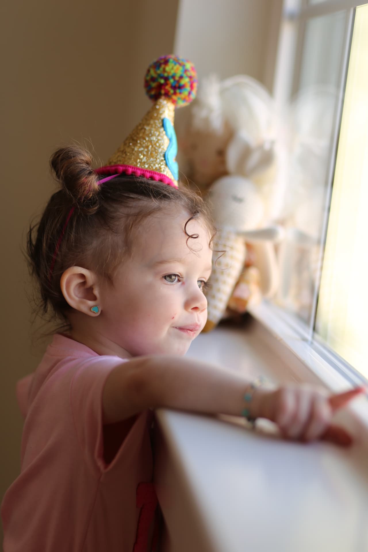 A girl in a glittery party hat leaning on the windowsill, smiling out.