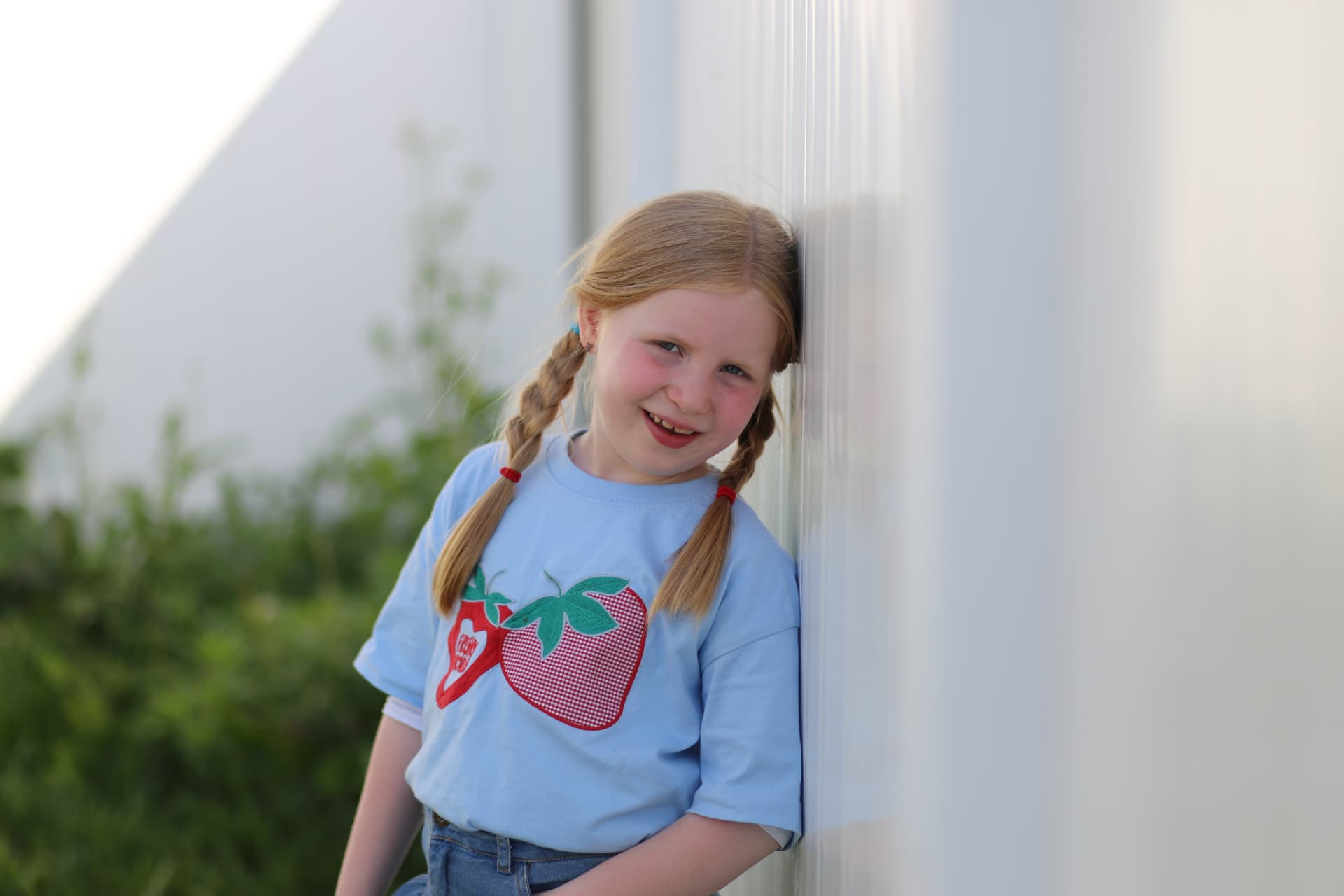 A girl with two braids grinning by the white wall in her strawberry t-shirt.