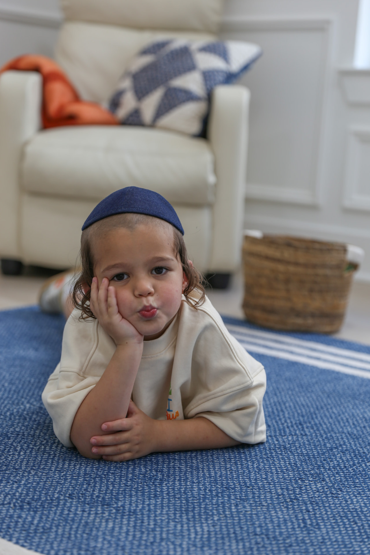 A boy in a kippah on the rug, lips puckered, cheek on his hand.
