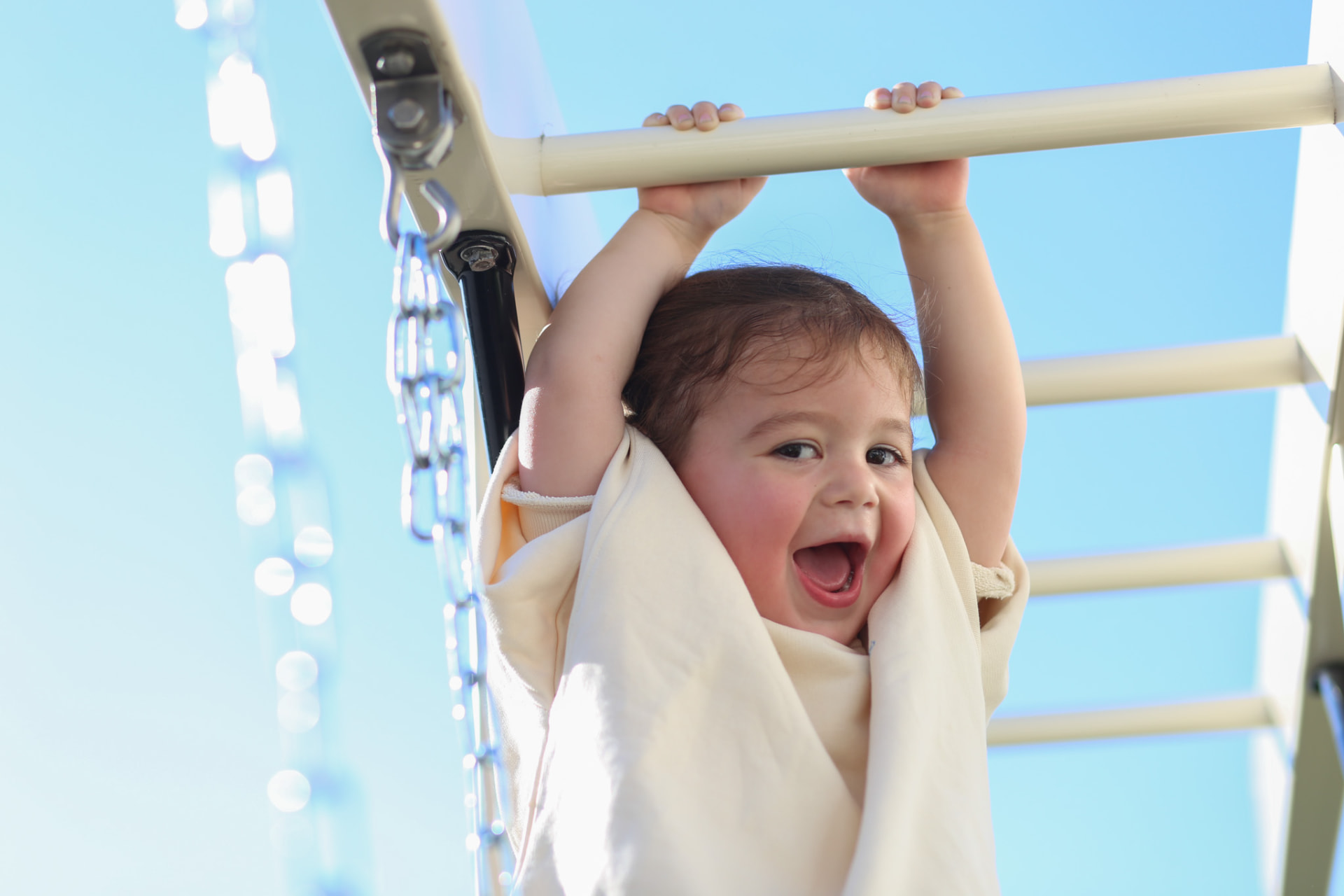 A baby hanging from the playset bar, laughing.