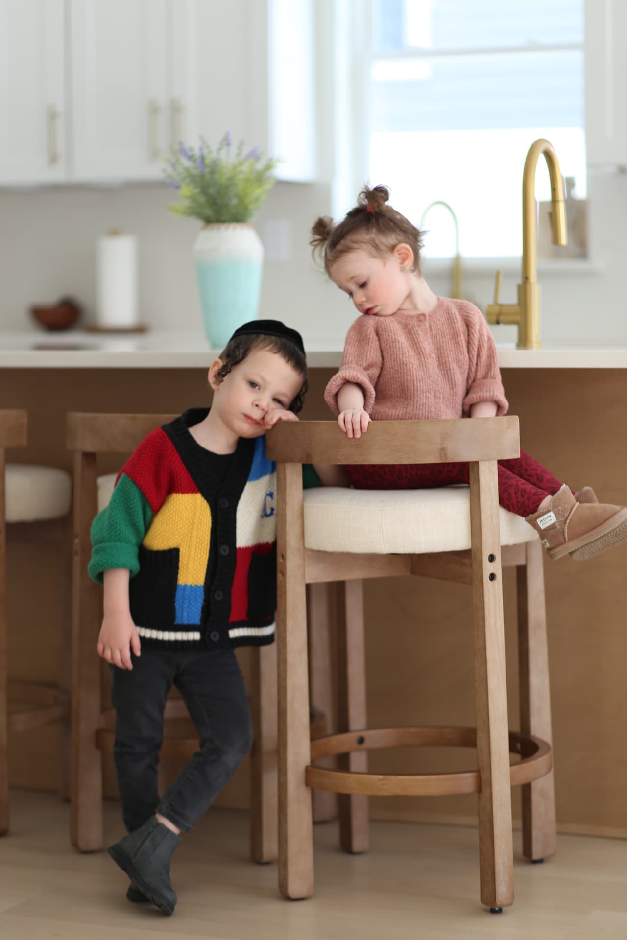 A brother leaning on a stool while his sister perches on top in the kitchen.