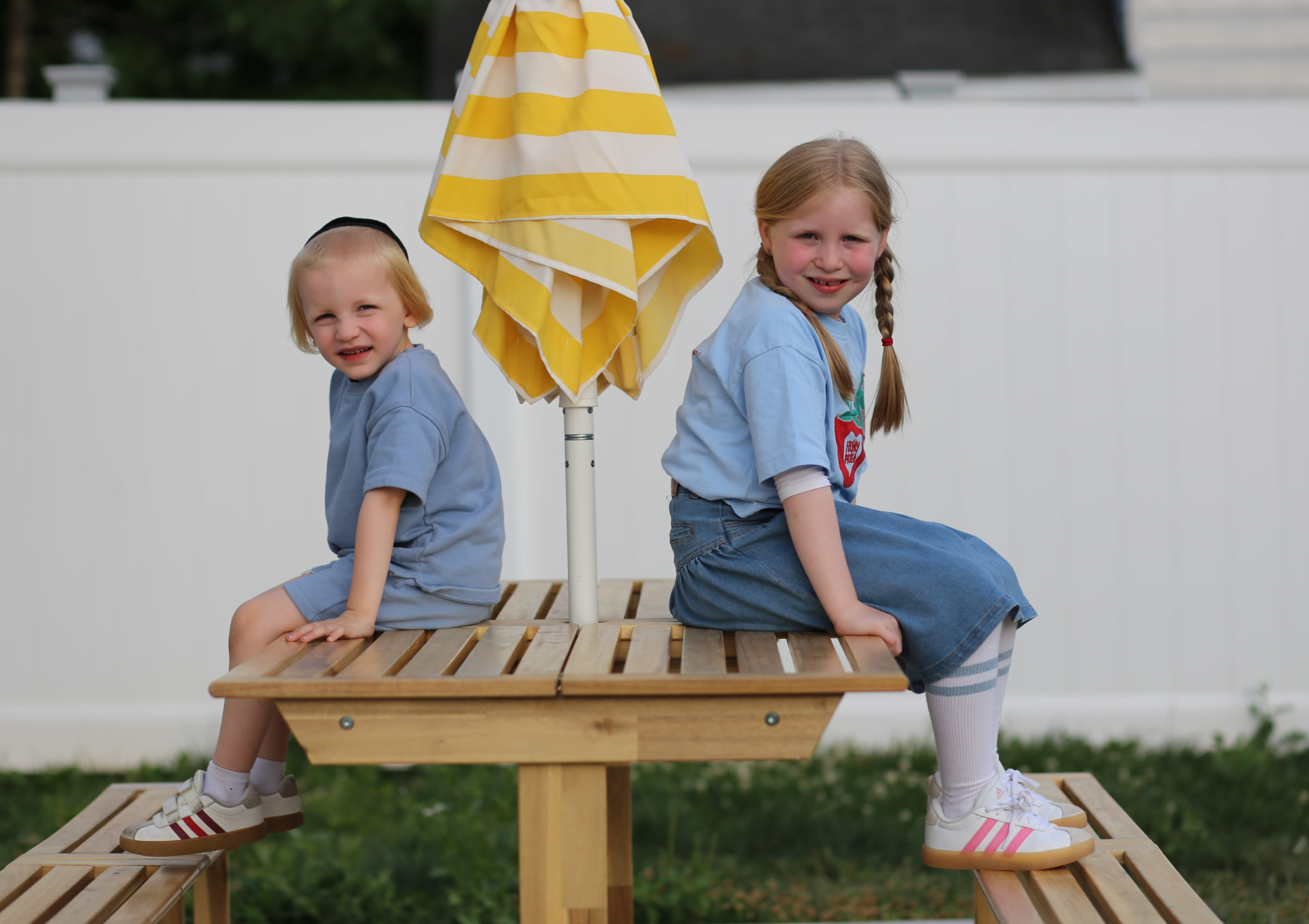 Two kids sitting side by side on the picnic table, the striped umbrella between them.