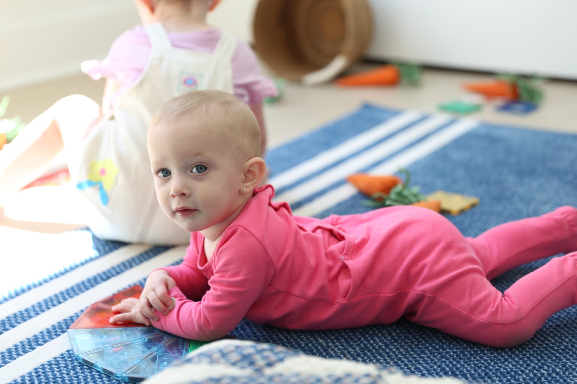 A baby in pink lying on the striped rug, looking up while her sister plays.